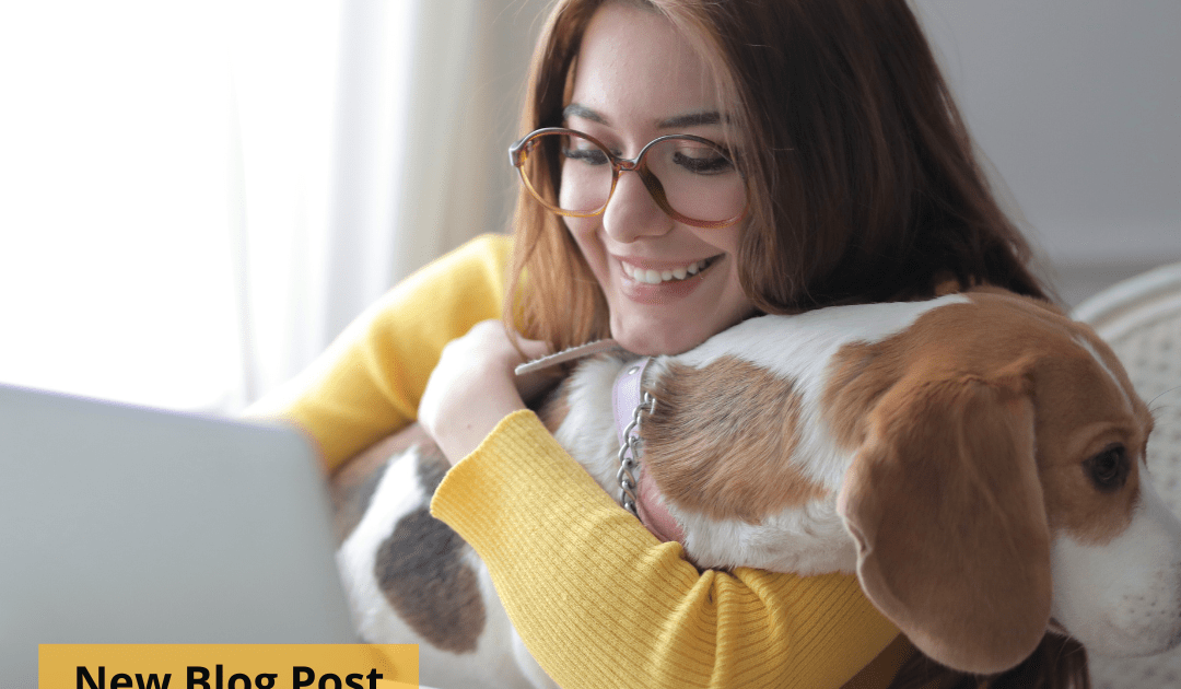 Woman wearing glasses and holding a dog sits at a computer.
