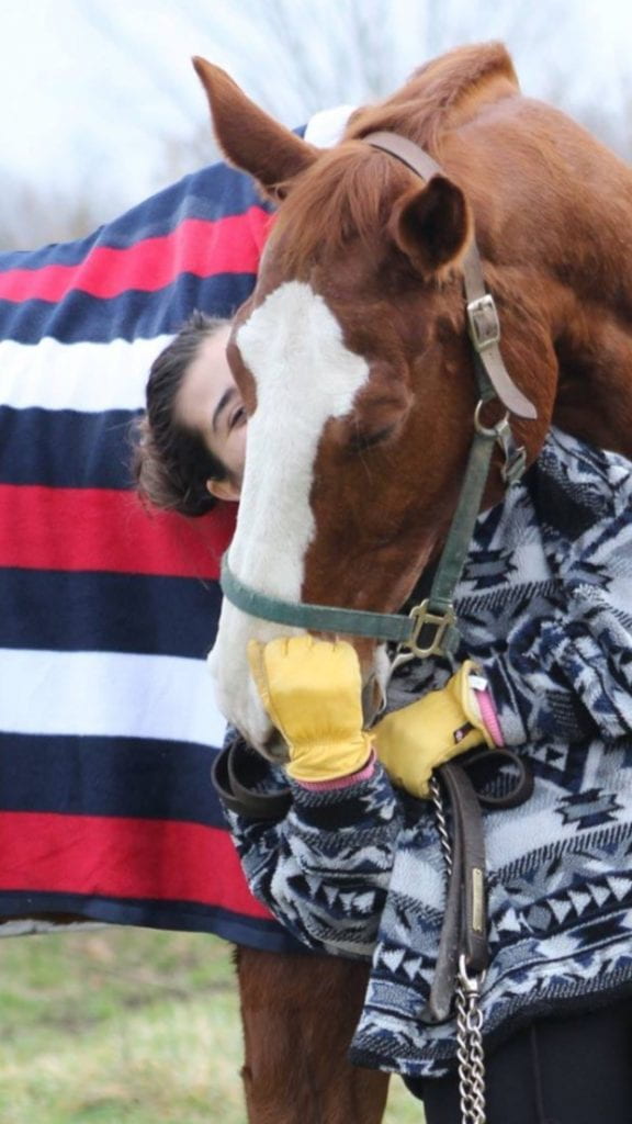 Cameron posing with Earl, a brown horse with a white stripe down his nose. Earl has a navy, red and white striped blanket on. 
